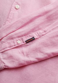 Camicia di lino rosa chiaro con bottoni bianchi, dotata di un'etichetta con la scritta "SUPERDRY". Il tessuto ha una consistenza morbida e dettagli di cucitura visibili.