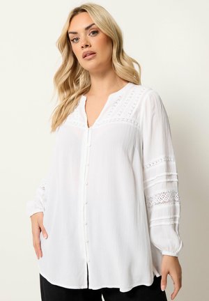 Donna bionda che indossa una camicetta bianca ampia con bottoni, dettagli in pizzo e pieghe sulle maniche, in piedi davanti a uno sfondo chiaro e uniforme.