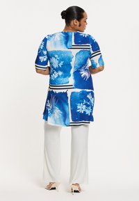 Blaue Tunika mit Blumenmuster und geometrischen Mustern. Kurze Ärmel, abgerundeter Saum. Kombiniert mit weißen, weiten Hosen und hellen Absätzen.