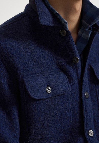 Marineblaue Wolljacke mit einem geknöpften Kragen, zwei Brusttaschen mit Klappen und Metallknöpfen mit dekorativem Emblem. Strukturierter Stoff.