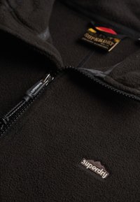 Zwarte fleece jas met rits en een zachte textuur, voorzien van Superdry-logo op de borst en een merkbare ritstrekker.