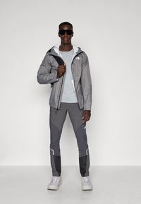 Pilka neperšlampama striukė su gobtuvu, šviesiai pilku vidiniu sluoksniu, derinama su pilkomis kelnėmis, kurias puošia tamsiai pilkos detalės ir atspindinčios juostos, dėvima su akiniais nuo saulės.