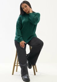 Gruba zielona sweterka z dzianiny z długimi rękawami, w połączeniu z czarnymi spodniami o szerokich nogawkach i czarnymi botkami za kostkę, siedząca na drewnianym stołku.