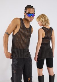 Schwarze Netz-Tanktops verfügen über ein lockeres, offenes Strickdesign. Das männliche Modell kombiniert es mit lässigen Jeans; das weibliche Modell trägt schwarze Shorts.