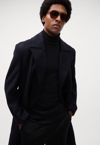 Zwarte wollen overjas met brede revers, gedragen over een zwarte coltrui. Model draagt donkere zonnebrillen, gestyled met natuurlijke krullend haar.