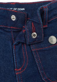 Tela de mezclilla azul oscuro con costuras rojas en contraste y herrajes plateados. Cuenta con un cierre de botón y pasadores para cinturón en la cintura.