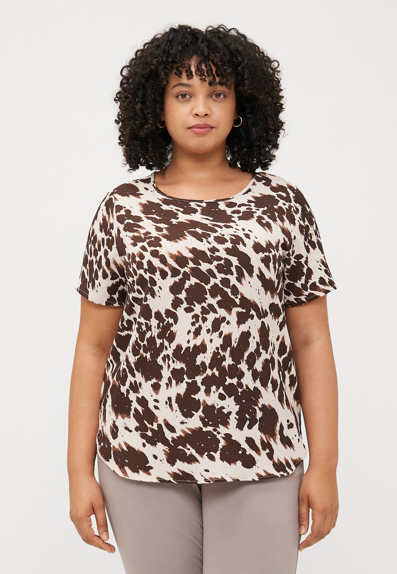 Curly-haired woman wearing a patterned brown and beige t-shirt, standing against a plain white background, looking directly at the viewer.
