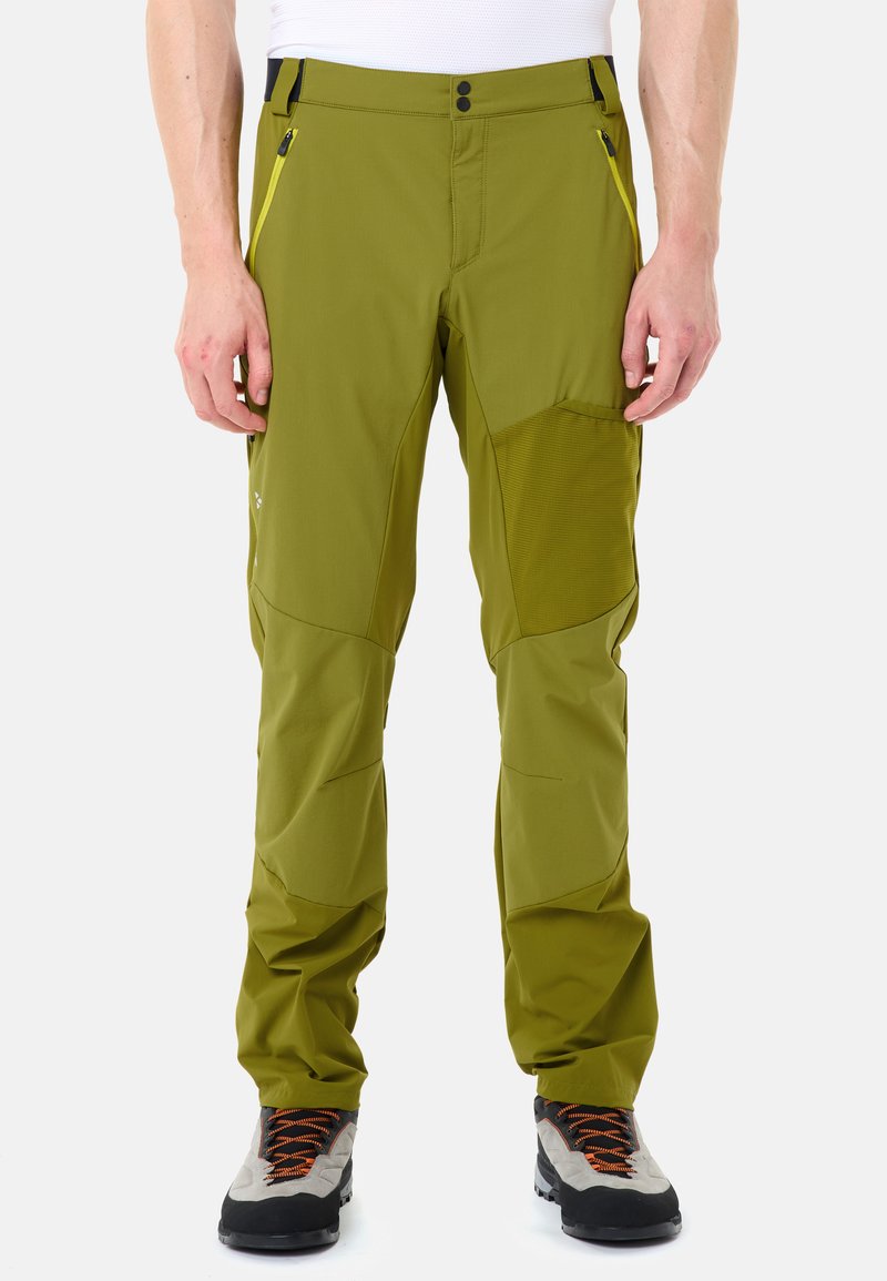 Homme portant un pantalon de plein air vert olive avec des poches latérales zippées et des chaussures de randonnée noires et grises avec des lacets orange, debout sur un fond blanc.