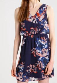 Robe à imprimé floral avec un décolleté en V profond, une conception sans manches, une taille élastique et une jupe en A fluide dans des teintes marine, rouge et rose clair.