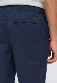 Homme portant un pantalon bleu marine avec une poche passepoilée à l'arrière et une petite étiquette noire indiquant "Solid Denmark Since 1999," main détendue sur le côté.