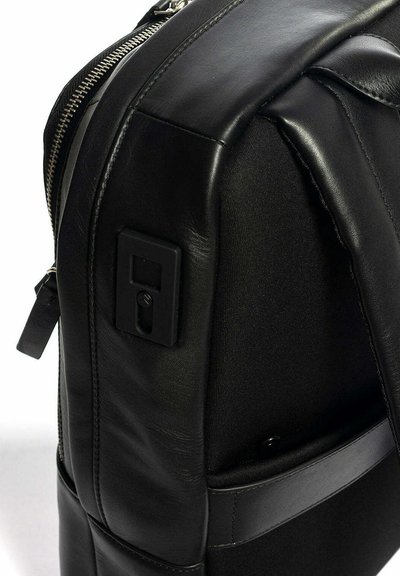 Zaino nero realizzato con una combinazione di pelle liscia e tessuto testurizzato, dotato di una cerniera argentata, una tasca laterale e un dettaglio in metallo rettangolare.