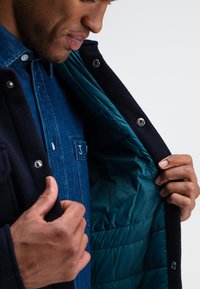 Navy outer jacket with a soft texture, shown unbuttoned to reveal a smooth turquoise lining. Layered over a blue denim shirt with a pocket.