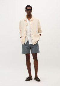 Jachetă bej cu nasturi și buzunar, cămașă albă, pantaloni scurți gri tip cargo și pantofi bruni. Model cu ochelari de soare închiși la culoare. Design simplu.