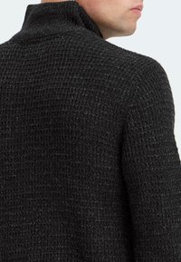 Kulsort strikket sweater med høj krave, der har et tekstureret mønster og en afslappet pasform. Materialet ser varmt og hyggeligt ud.