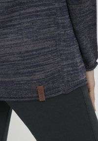 Tummansininen teksturoitu neulepaita, jossa on ruskea merkitarran hemlä, yhdistettynä tumman vihreisiin kapeisiin housuihin. Painotus kankaan ja värien kontrastiin.