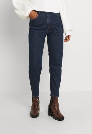 Jeans de mezclilla oscura de tiro alto con un corte ajustado y cinco bolsillos, combinados con botas de tobillo brillantes de color marrón con un tacón grueso.