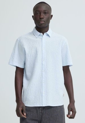 Modèle masculin à la peau foncée portant une chemise à manches courtes bleu clair texturée à boutons et un pantalon gris foncé, sur un fond uni.