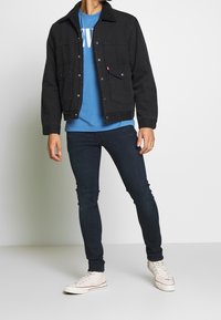 Black denim jacket with a sherpa collar, blue graphic t-shirt, dark jeans, and white sneakers. Features button closure and front pockets.