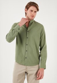Chemise à manches longues, verte olive, avec un col montant, fermeture à boutons et un tissu texturé. Présente des boutons noirs et un ourlet arrondi.