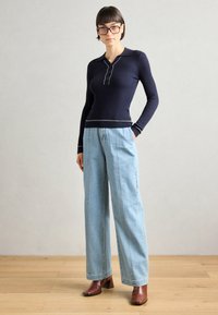 Top de manga longa em azul marinho com nervuras, com colarinho e fechamento de botões de pressão, combinado com jeans de perna larga em azul claro e sapatos de salto em castanho.