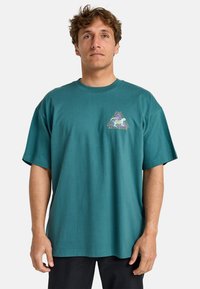 Teal cotton t-shirt with short sleeves, featuring a triangle graphic print in multiple colors on the left chest. Relaxed fit design.