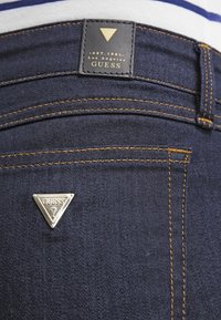 Vaqueros de mezclilla azul oscuro con costuras naranjas. Presentan una etiqueta con un logo triangular y un adorno de triángulo plateado con la inscripción "GUESS".