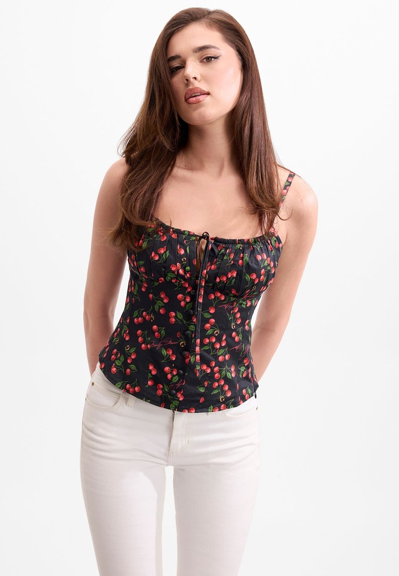 Femme aux longs cheveux bruns portant un débardeur noir à motifs cerises et un pantalon blanc, debout devant un fond blanc uni.