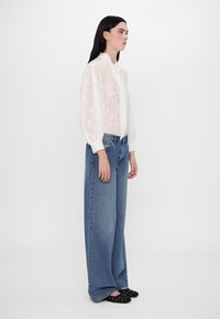 Blouse blanche transparente avec une texture florale et un col montant, associée à un jean bleu à taille haute et jambes larges, ainsi qu'à des chaussures à enfiler noires ornées.