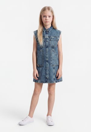 Ung pige iført en ærmeløs denimkjole med forlommer og hvide sneakers står på en ensfarvet hvid baggrund.