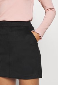 Svart mocka mini kjol med sidofickor, med en slät textur och hög midja. En rutig rosa långärmad topp syns delvis.