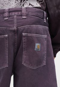 Μωβ παντελόνι από τζιν με πίσω τσέπες, με το λογότυπο της Carhartt στην δεξιά τσέπη, φορεμένο με εκτυπωμένο πουκάμισο και σκούρο μακρυμάνικο τοπ.