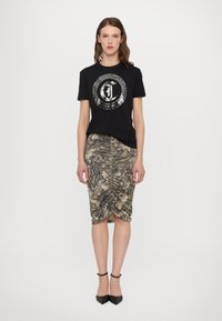 T-shirt en coton noir avec un motif métallique, associé à une jupe crayon froncée beige et marron à motifs, portée avec des talons noirs.