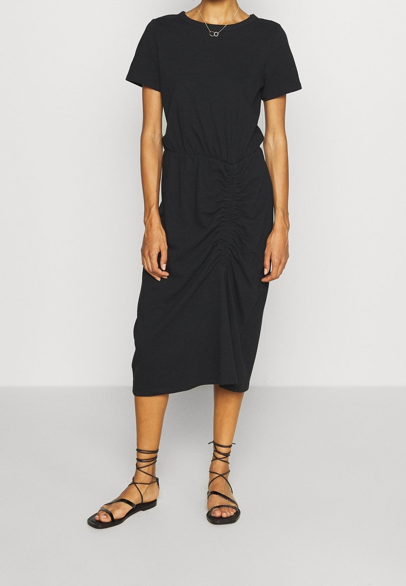 Robe midi noire à manches courtes avec des détails froncés à la taille, fabriquée en tissu doux. Associée à des sandales plates noires à brides.