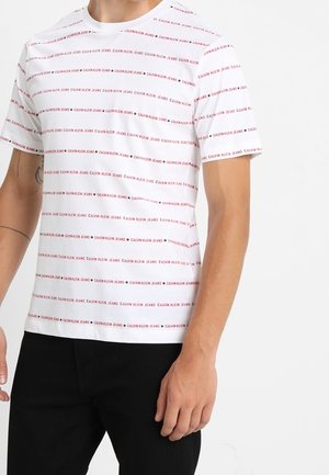 Biała koszulka z krótkim rękawem z czerwonymi poziomymi paskami z napisem "Calvin Klein Jeans", noszona przez osobę w czarnych spodniach z widocznymi tatuażami na ramionach.