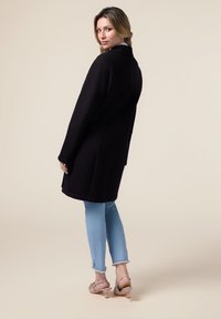Cappotto di lana nera con design strutturato, fessure laterali; abbinato a jeans azzurri chiari e tacchi nudo. Silhouette semplice ed elegante.
