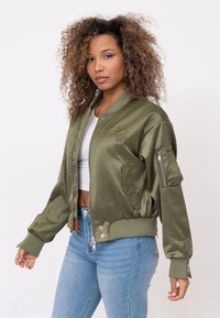 Olijfgroene bomberjack van satijn met een ribbelkraag, manchetten, zijzakken en geborduurde logo-details op de voorkant.