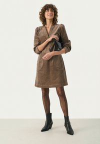 Ruskea corduroy-mekko, jossa on V-pääntie, pussimaiset hihat ja kaksi etutaskua, yhdistettynä mustiin korkokengät ankle boots -kenkiin ja pieneen mustaan käsilaukkuun.