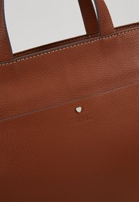 Sac fourre-tout en cuir brun texturé avec un accent cœur argenté et un logo en relief. Présente un design structuré avec des poignées doubles et des coutures contrastantes.