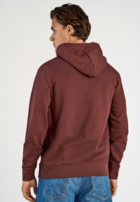 Felpa bordeaux con cappuccio regolabile, polsini e orlo a coste. Tessuto morbido, indossata con jeans in denim blu. Dettagli di cucitura visibili.