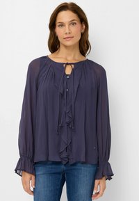 Blusa blu scuro realizzata in tessuto leggero, con una parte anteriore a volant, chiusura a fiocco al collo e maniche svasate. Semplici dettagli con bottoni.