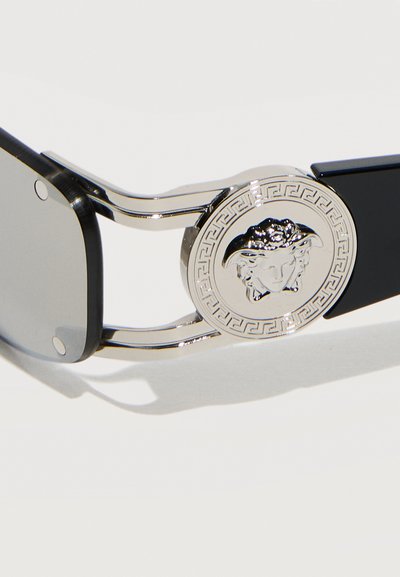 Πλαινό μέρος γυαλιών ηλίου με μαύρο σκελετό, ασημί έμβλημα Μέδουσας ανάγλυφο σε κυκλικό μεταλλικό διακοσμητικό και ανακλαστικό γκρι φακό σε λευκό φόντο.