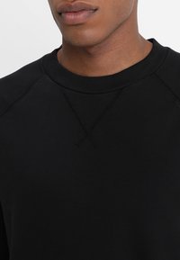 Černá mikina s vroubkovaným kulatým výstřihem, texturovaným švem a hladkým, jemným materiálem. Detailní pohled na límec a horní část hrudníku.