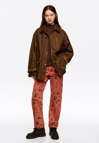 Rudas nepralaidus paltas su kelnių audiniu apykakle, vilkimas ant rudos bluzos. Rožinės kelnės su tamsiais dėmėtais raštais. Juodi bateliai su raišteliais.