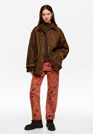 Chaqueta marrón impermeable con cuello de pana sobre una camiseta marrón. Pantalones rosas con patrones de salpicaduras oscuros. Botas negras con cordones.