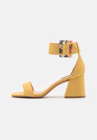 Keltaiset mokkasandaalit korolla, joissa on neliönmuotoinen solki, avonäinen malli ja paksu korko. Omaavat sileän pinnan ja minimalistisen tyylin.