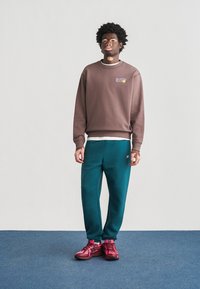 Καφέ φούτερ με μικρό λογότυπο, πετρόλ φόρμα και ροζ αθλητικά παπούτσια. Το σύνολο έχει άνετη γραμμή και μαλακές υφές.