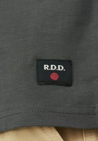 Tessuto grigio scuro con un'etichetta cucita che presenta "R.D.D." in testo bianco e un cerchio rosso su uno sfondo nero con linee diagonali.