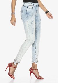 Jasnoniebieskie dżinsy z efektem postarzania oraz zdobieniami na nogawkach, zestawione z czerwonymi sandałami na wysokim obcasie z paskami.