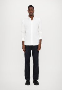 Camisa branca de manga longa com colarinho abotoado, combinada com jeans de ganga azul escuro e sapatos pretos. Design simples e clássico.