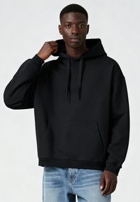 Fekete kapucnis pulóver elülső zsebbel, bordázott mandzsettákkal és laza szabással, világoskék, kopott hatású farmernadrággal párosítva.